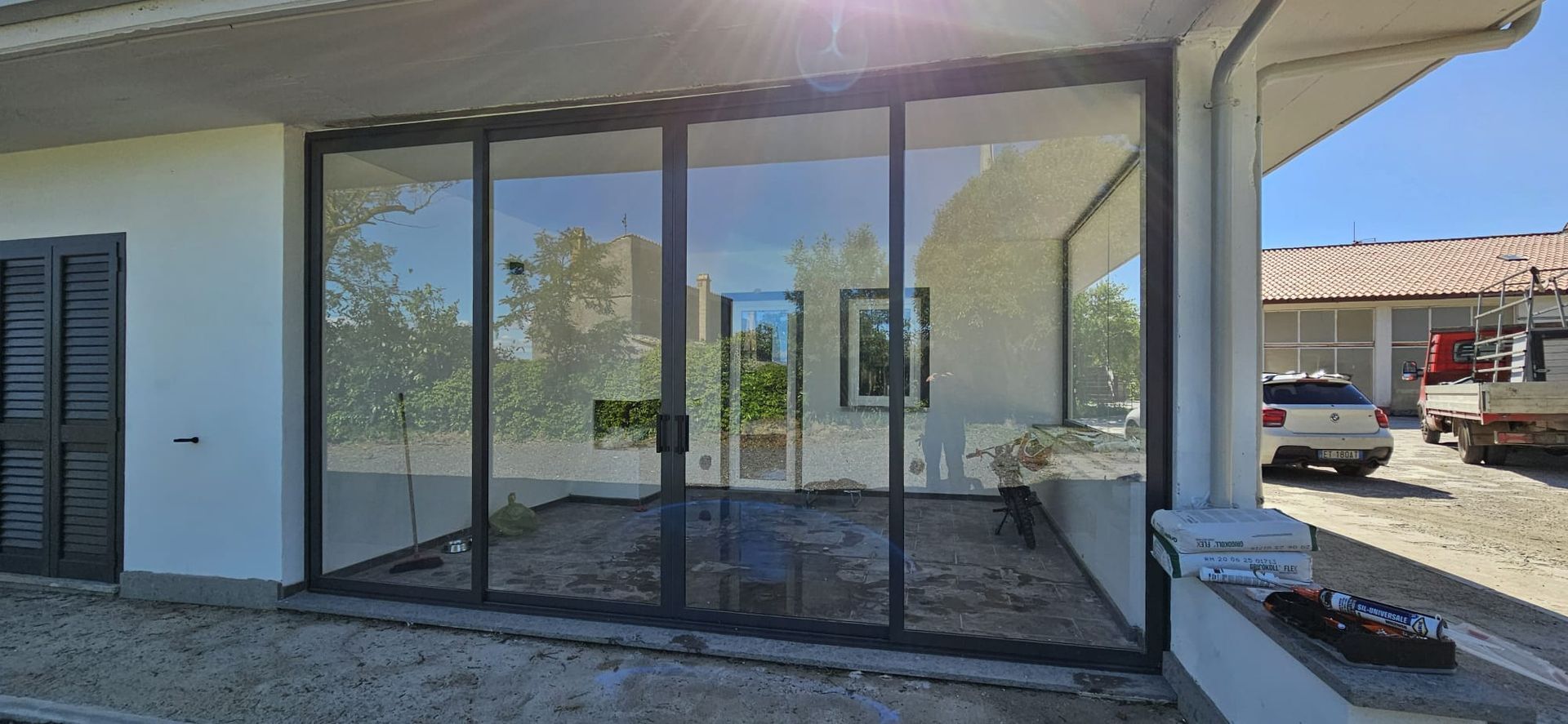 Vista esterna di un edificio con ampie vetrate che riflettono gli alberi e il cielo terso. Un'auto è parcheggiata di lato.
