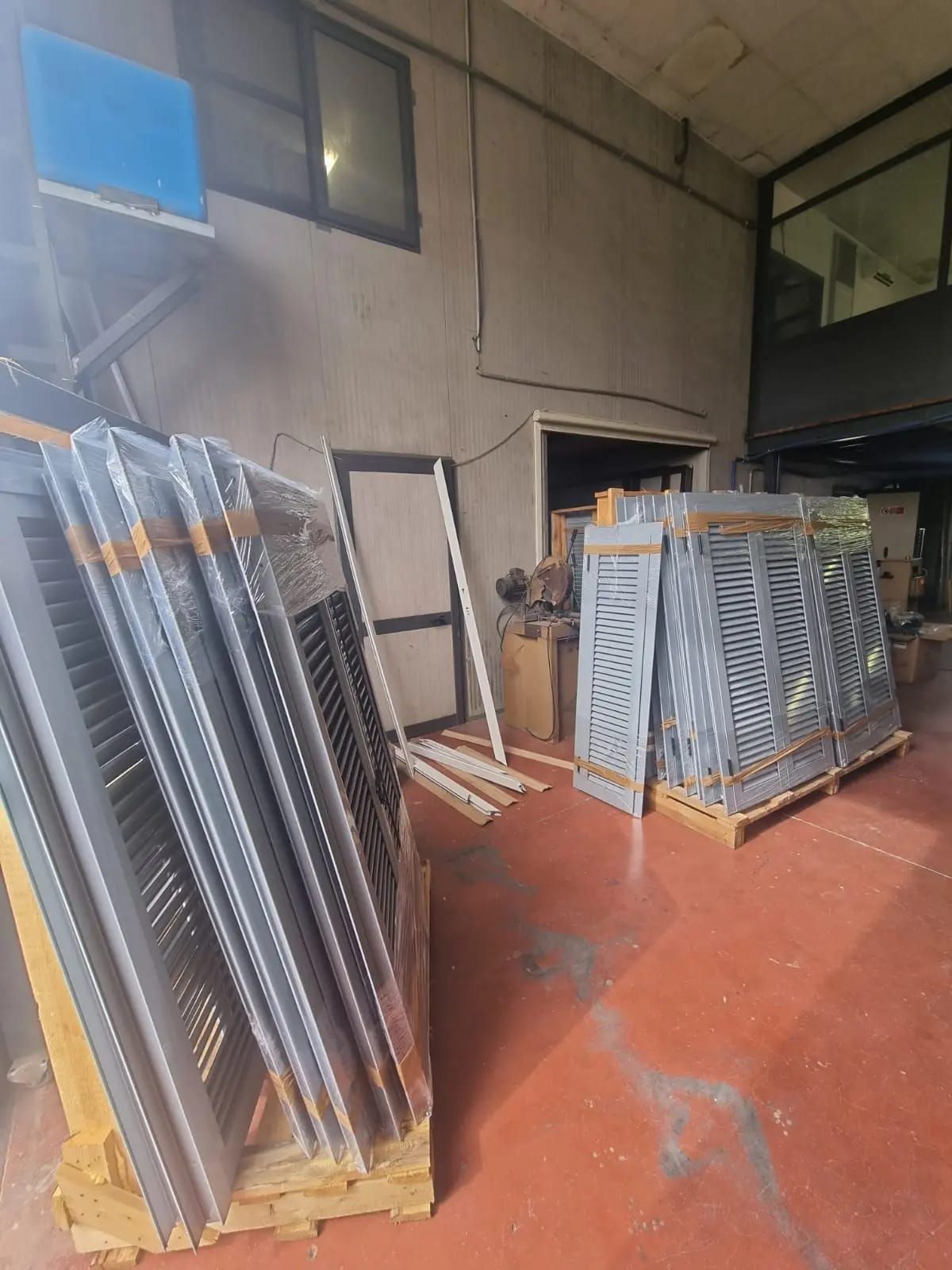 Pile di finestre in metallo grigi su pallet all'interno di un magazzino. Pavimento rosso, pareti neutre, luce naturale.