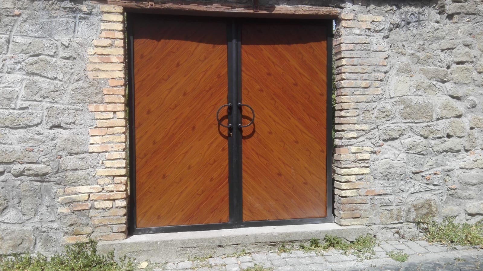 Doppie porte effetto legno marrone con maniglie nere, inserite in un muro di pietra con bordo in mattoni.