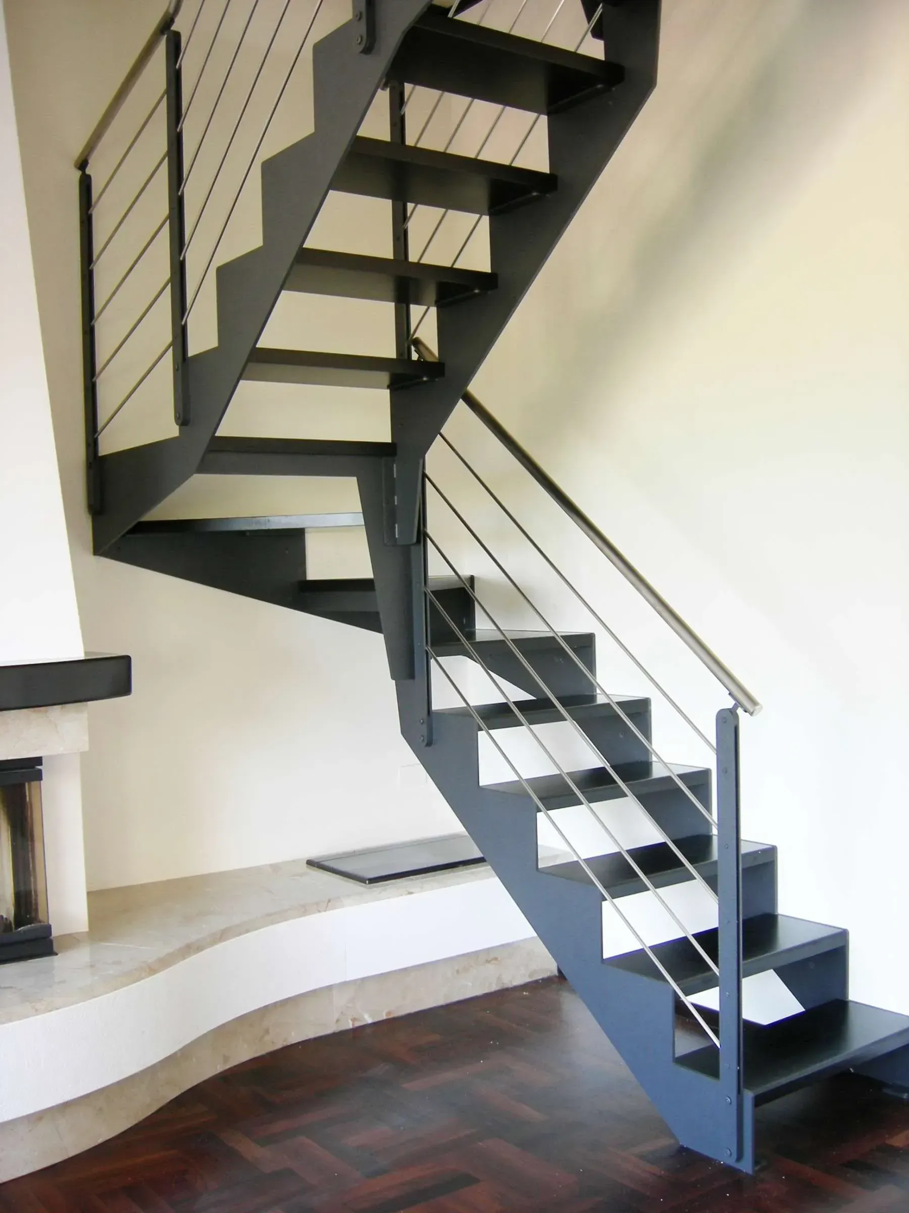 Moderna scala sospesa con struttura in metallo grigio, gradini neri e ringhiere in cavi metallici.