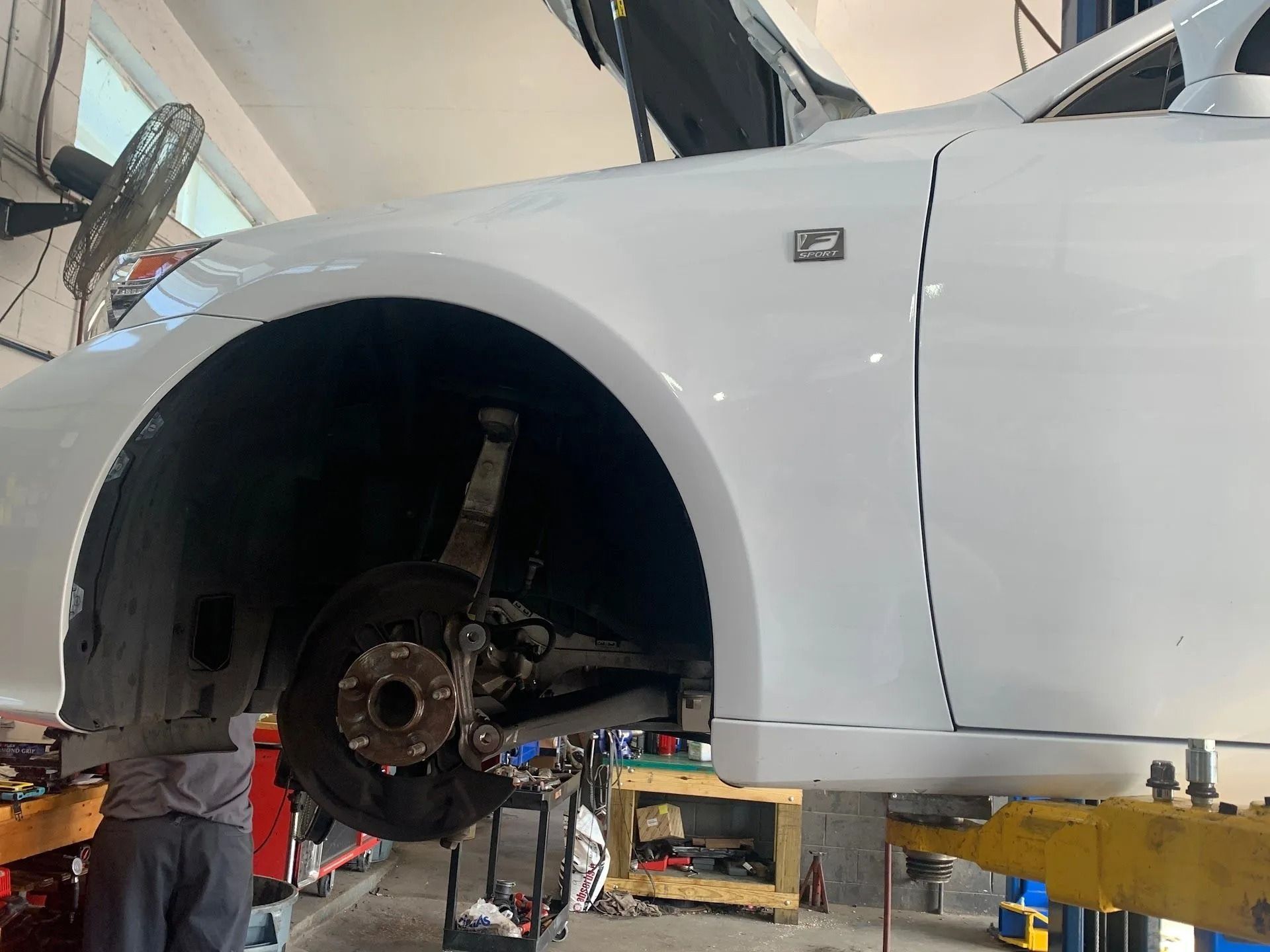 White car on a lift in a garage; front wheel removed, mechanic working.