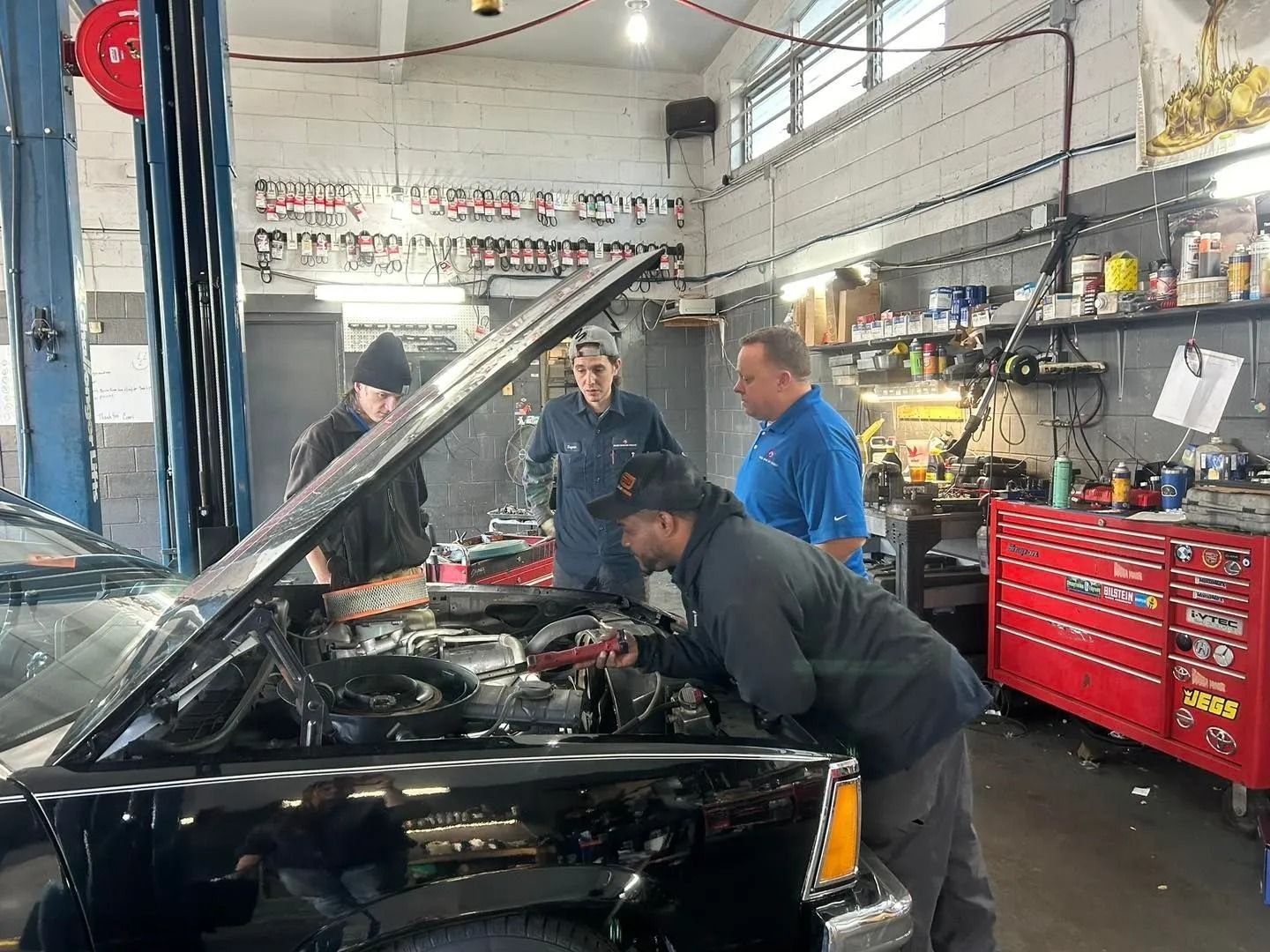 Mechanics examine a car engine in a garage. Three people are looking under the hood. Red tool cabinet in the background.