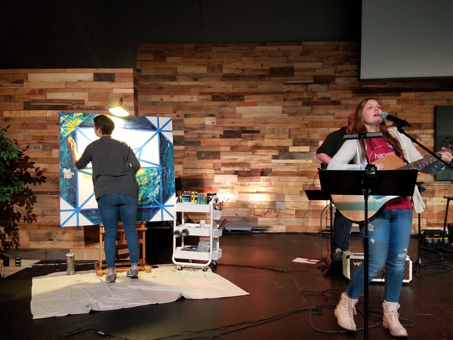 A woman is singing into a microphone while another woman paints a picture behind her.
