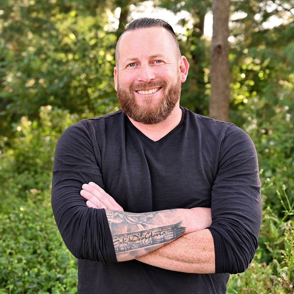 A man with a beard and tattoos is standing with his arms crossed and smiling.