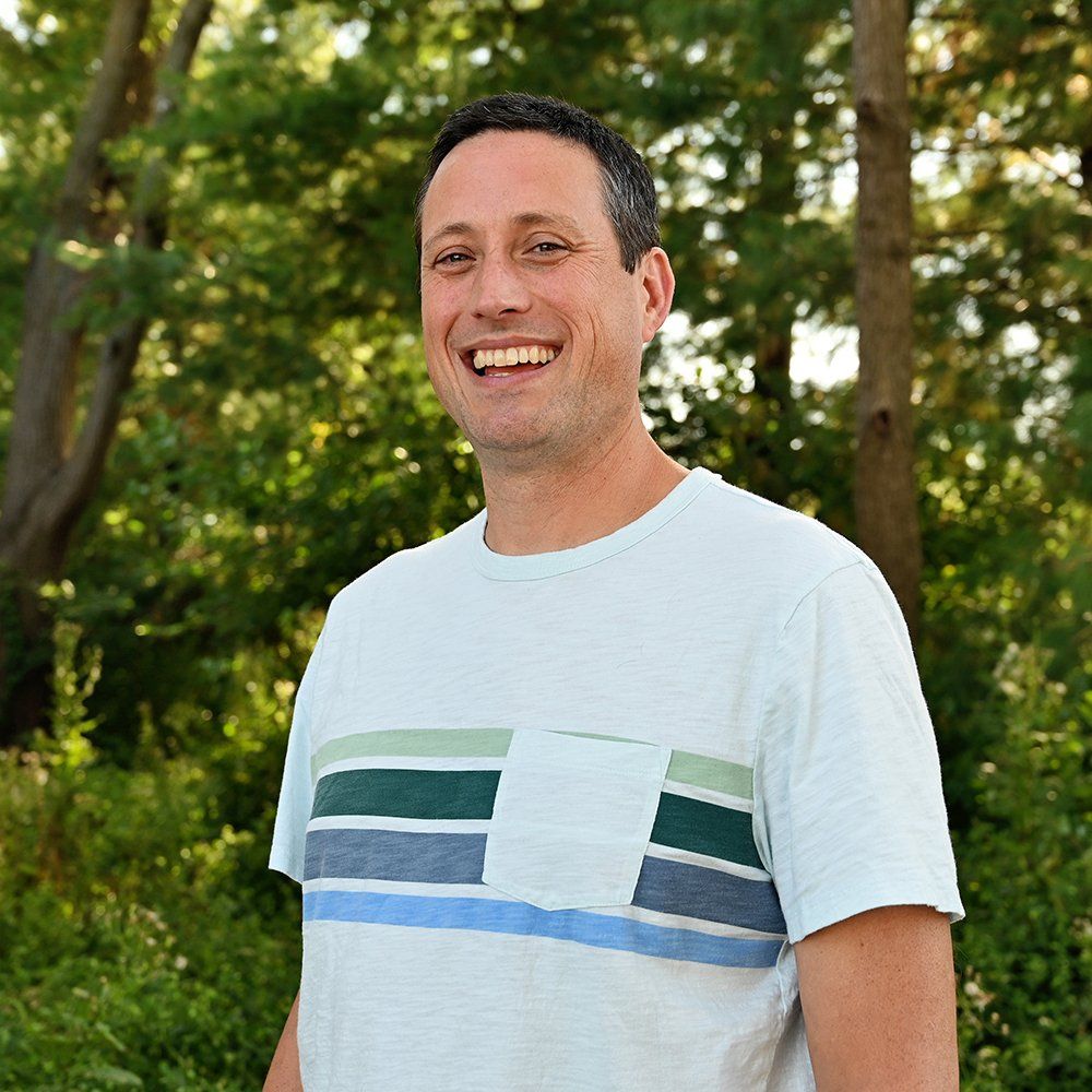 A man in a striped shirt is smiling in front of trees.