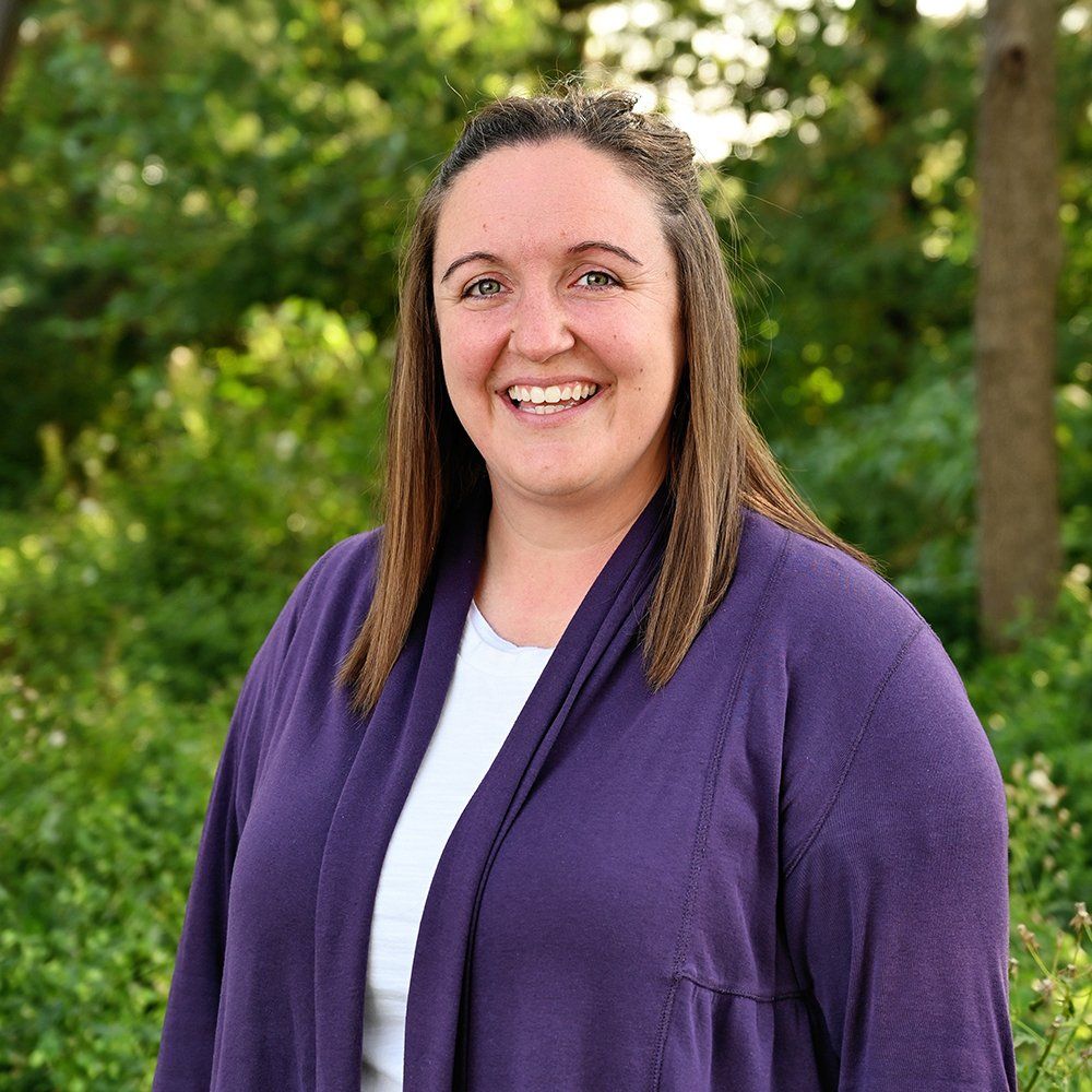 A woman wearing a purple cardigan and a white shirt is smiling for the camera.