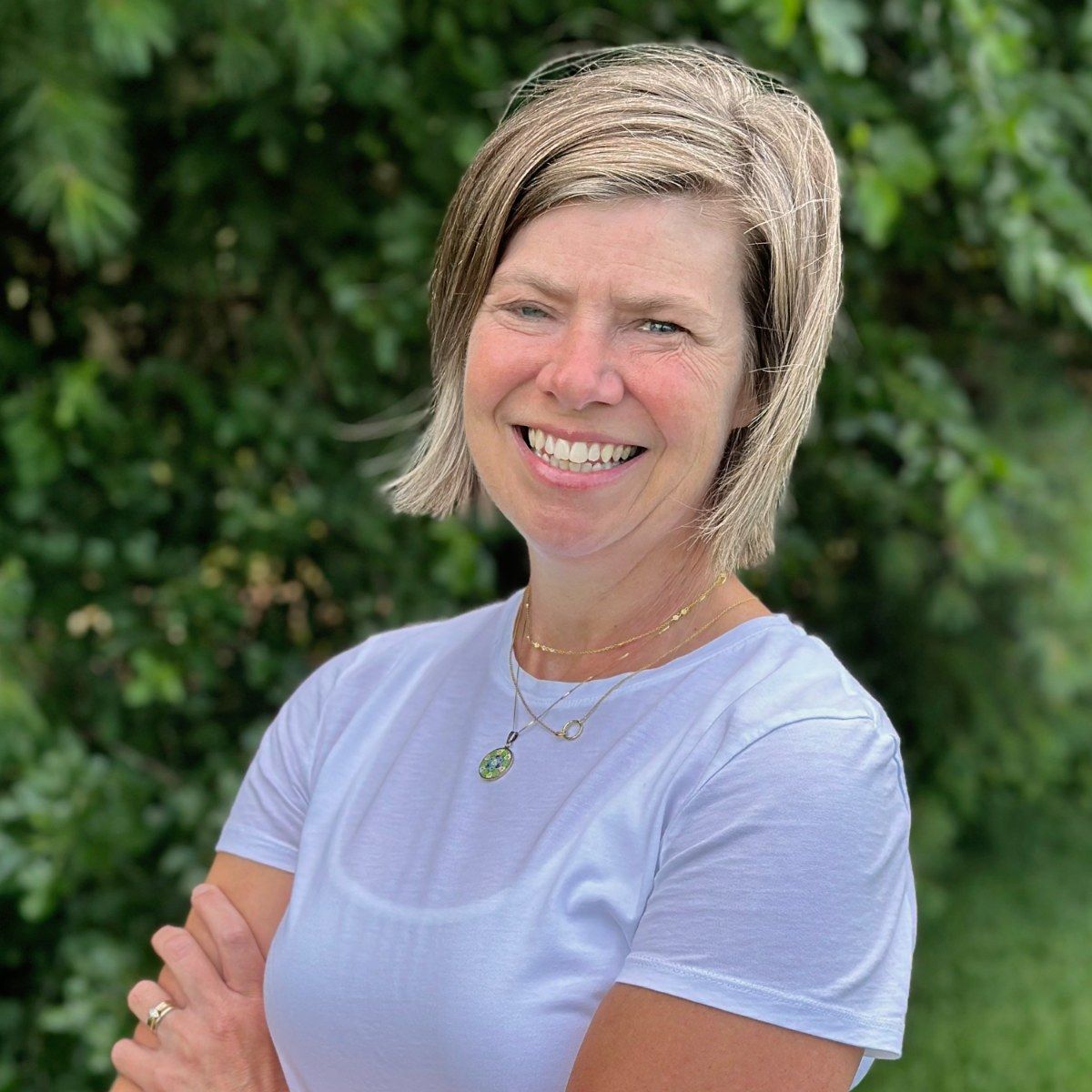 A woman wearing glasses and a green sweater is standing in front of a forest.