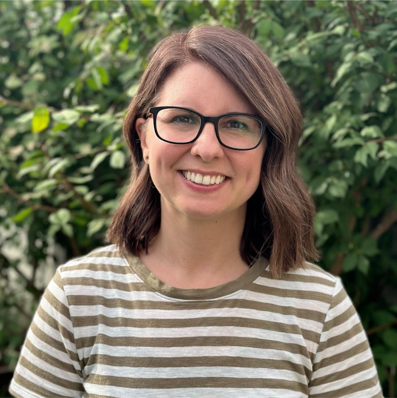 A woman wearing glasses and a green jacket is standing with her arms crossed and smiling.