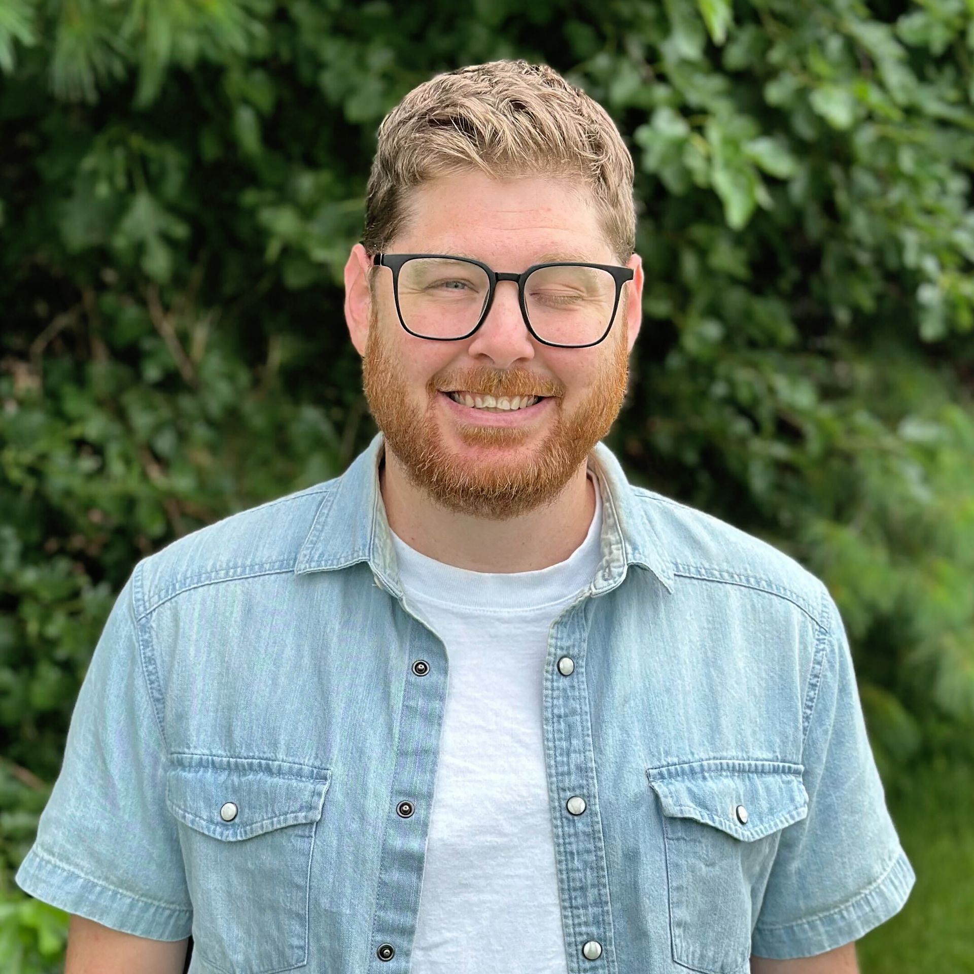 A man with a beard and glasses is smiling in front of a tree.