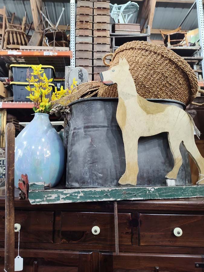 Wooden dog cutout next to metal bucket and woven basket on a wooden surface, with a blue vase and drawers.