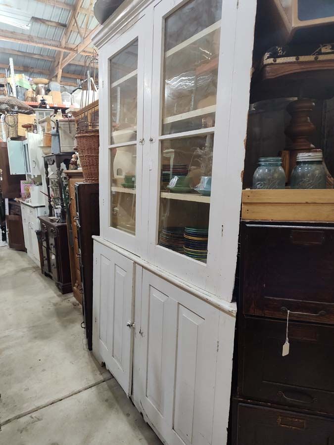 White antique cabinet with glass doors, filled with dishes, in a cluttered warehouse setting.