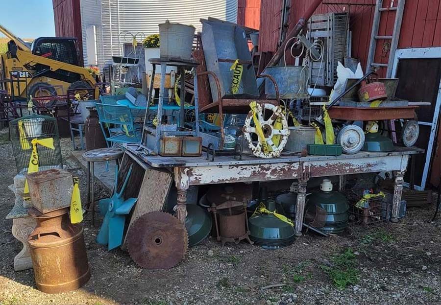 A cluttered outdoor display of rusty metal objects and tools, including tables, buckets, and wheels, against a red barn.