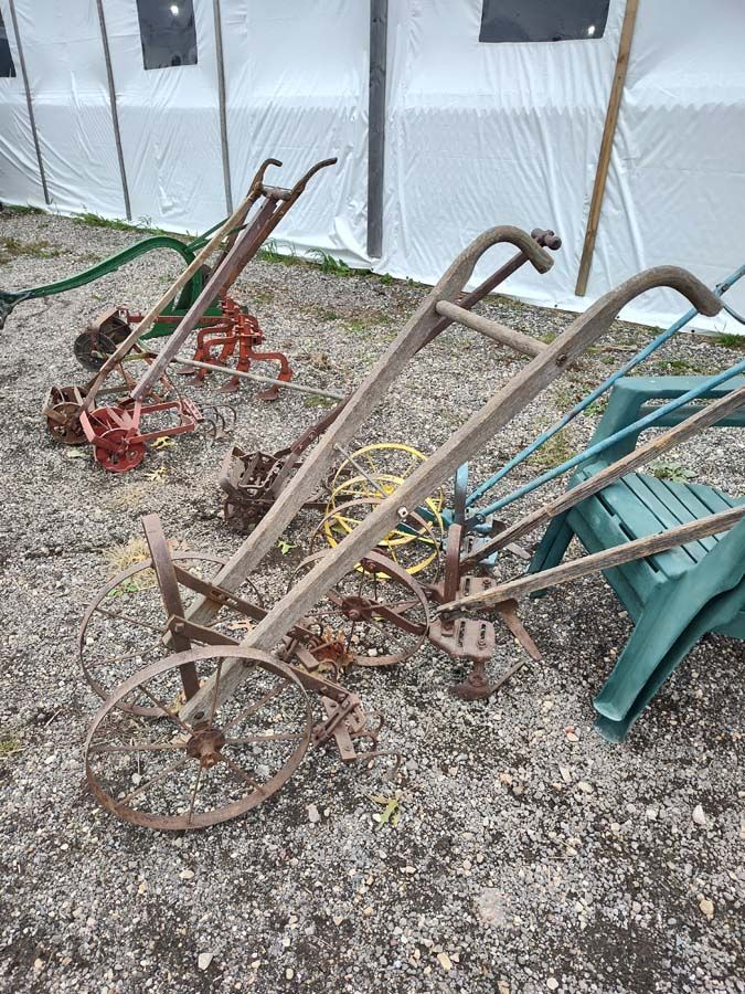 Three antique gardening plows on gravel, beside a green plastic chair and white sheet tent.