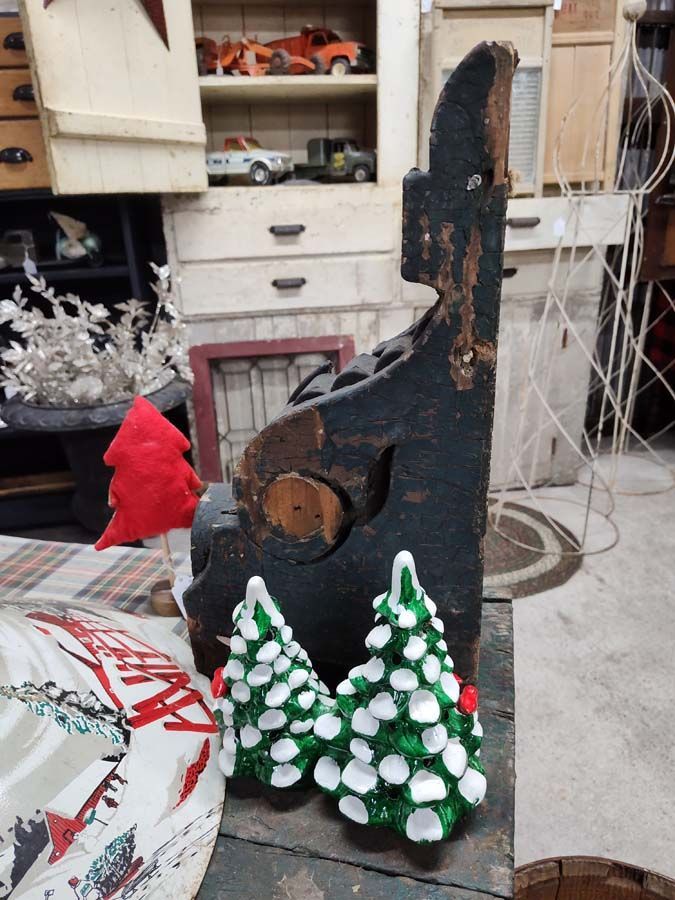 Black, weathered metal art with two small Christmas tree figurines. A red tree and fabric visible.