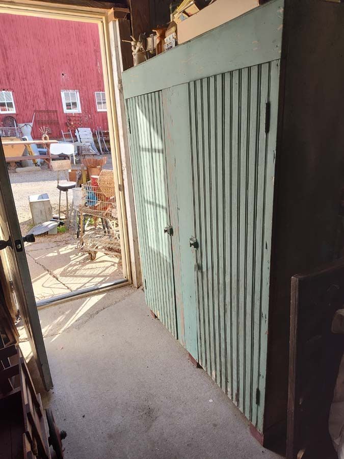 Tall, ribbed, teal-painted cabinet with two doors in a cluttered garage. Red-roofed building visible outside.