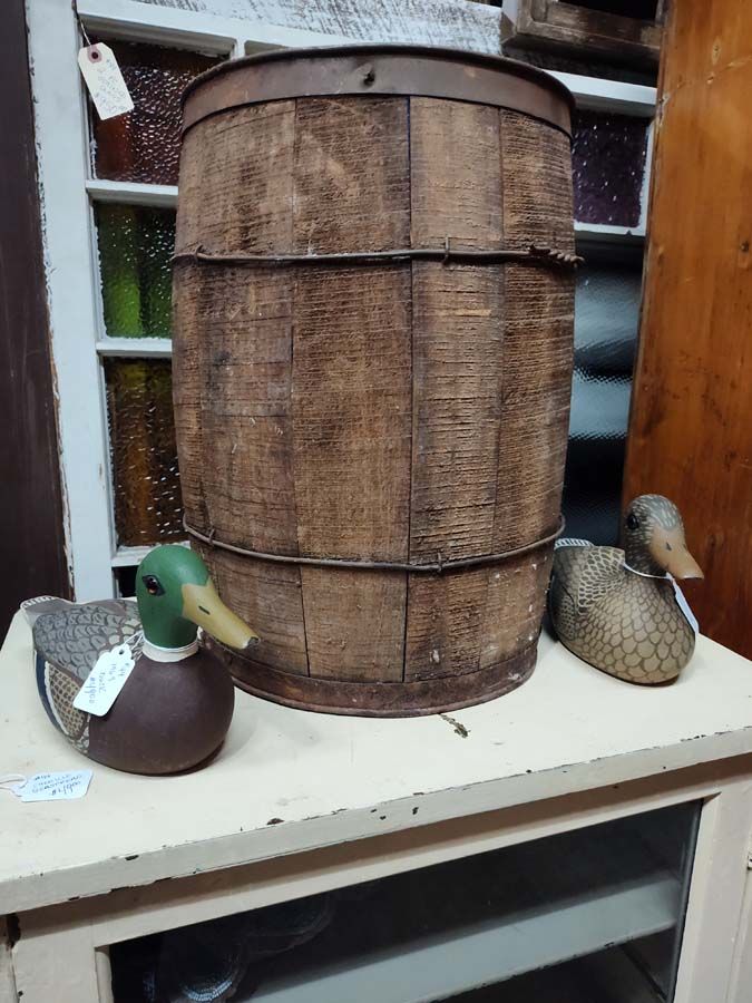 Wooden barrel with metal bands, flanked by two duck decoys, on a white shelf, in a shop.