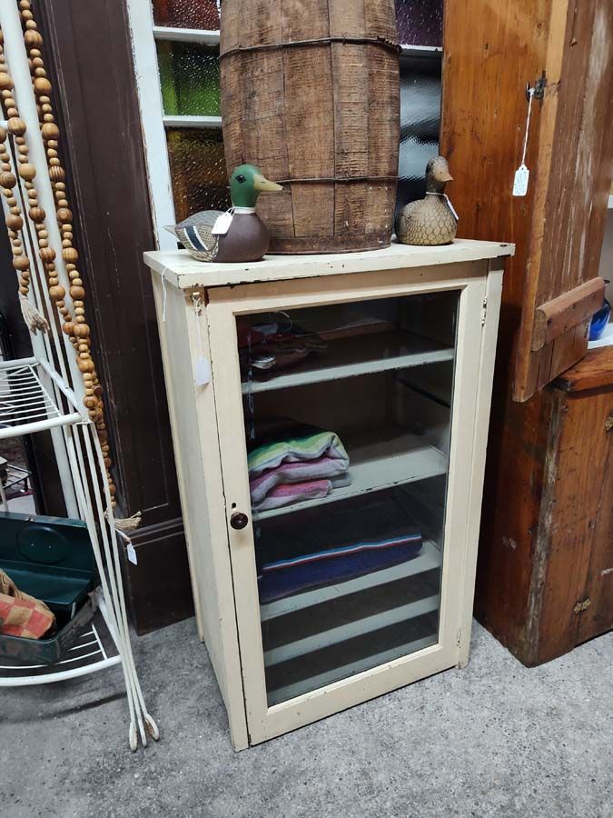 Cream-colored cabinet with glass door. Inside are shelves with folded textiles. A barrel and duck decoys sit on top.
