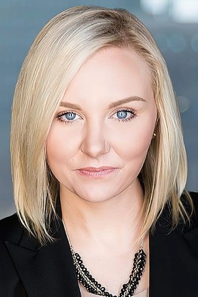 A woman with blonde hair and blue eyes is wearing a black jacket and a black necklace.