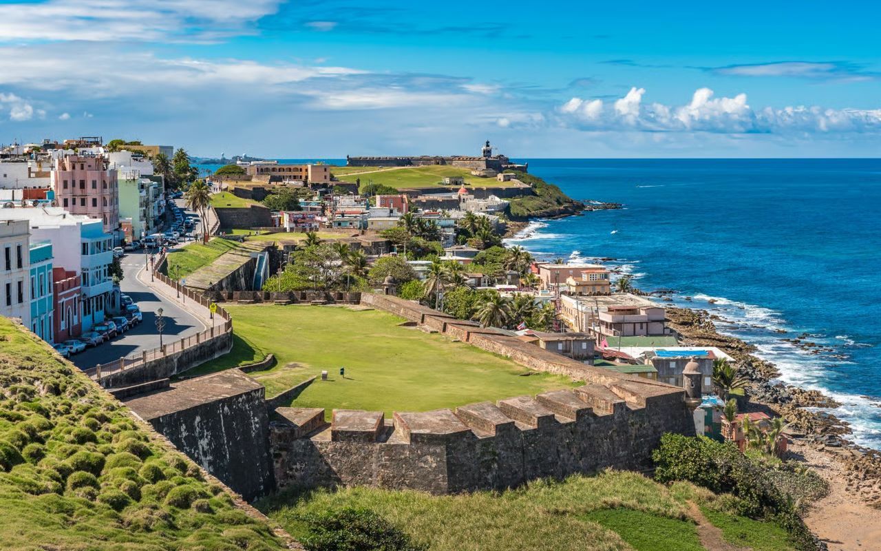 Coastal view of Old San Juan, Puerto Rico, with fortress walls, buildings, and the Atlantic Ocean under a blue sky.