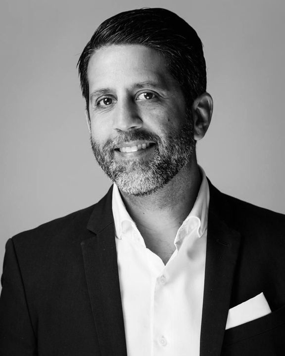 Josean in a blazer and open-collared shirt, smiling with a pocket square, studio shot.