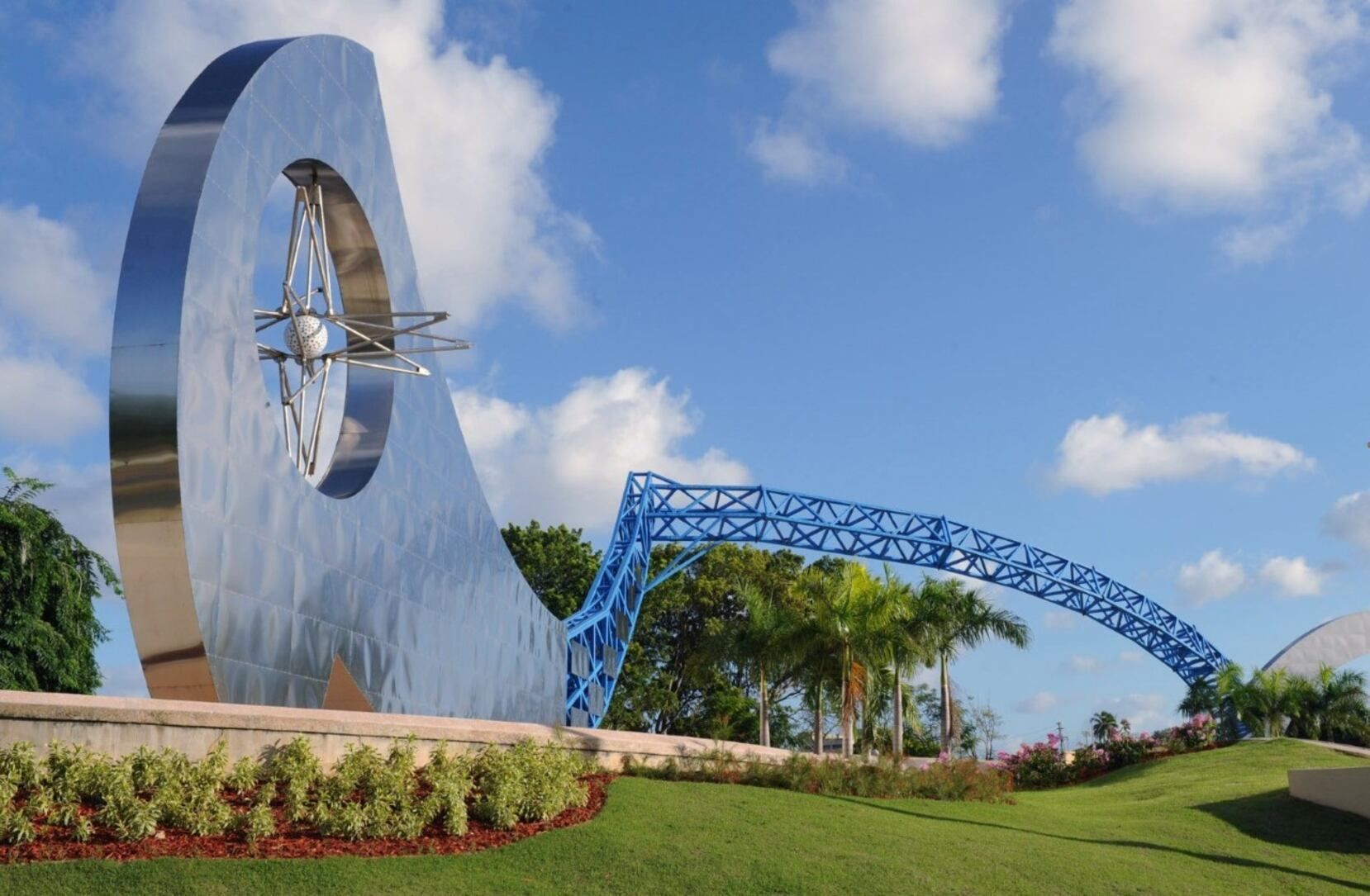 Large metallic sculpture, with blue archway, set in a grassy area under a blue sky with clouds.
