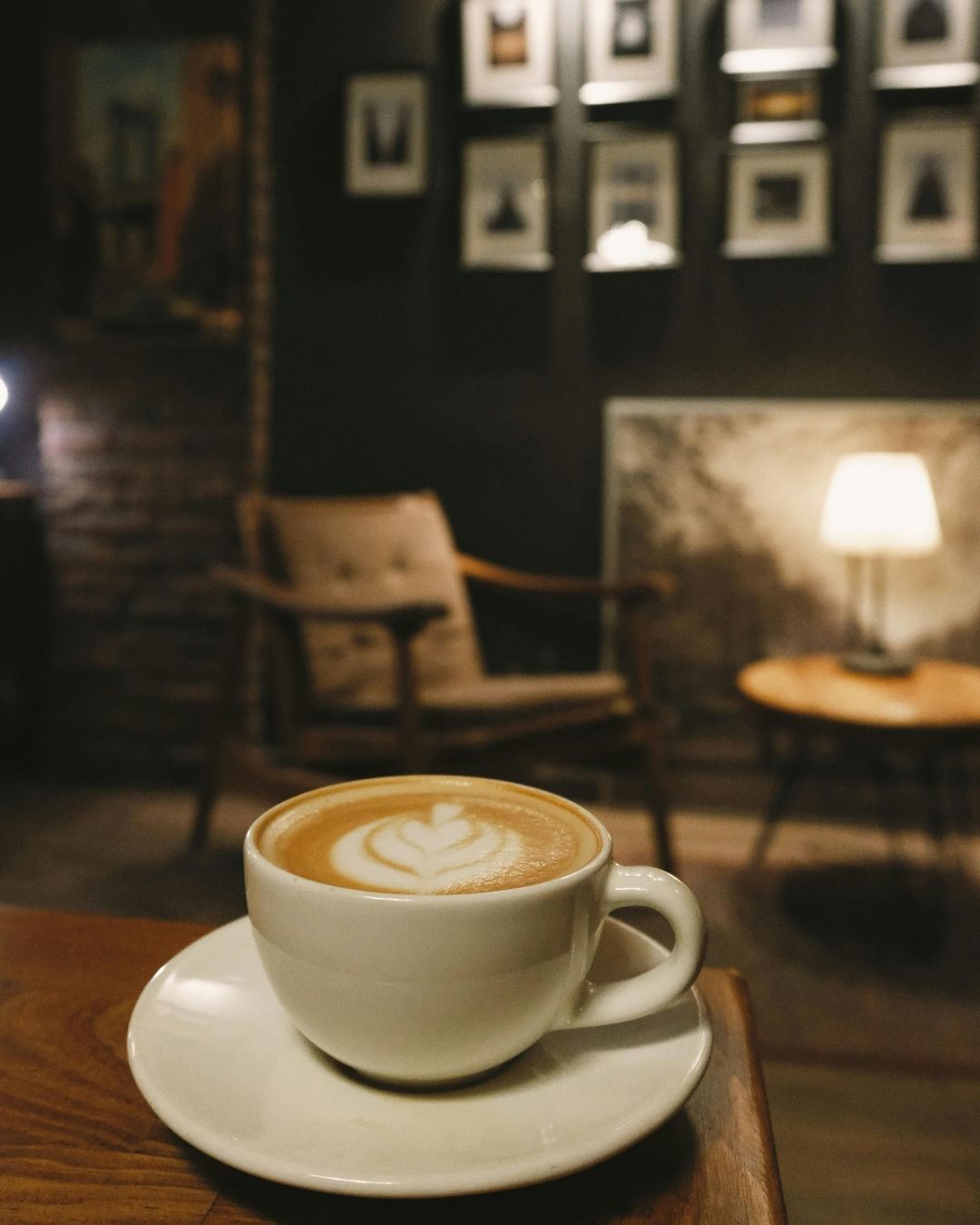 A white cup of latte with heart-shaped foam art sits on a wooden table in a dimly lit, cozy cafe with framed wall art.