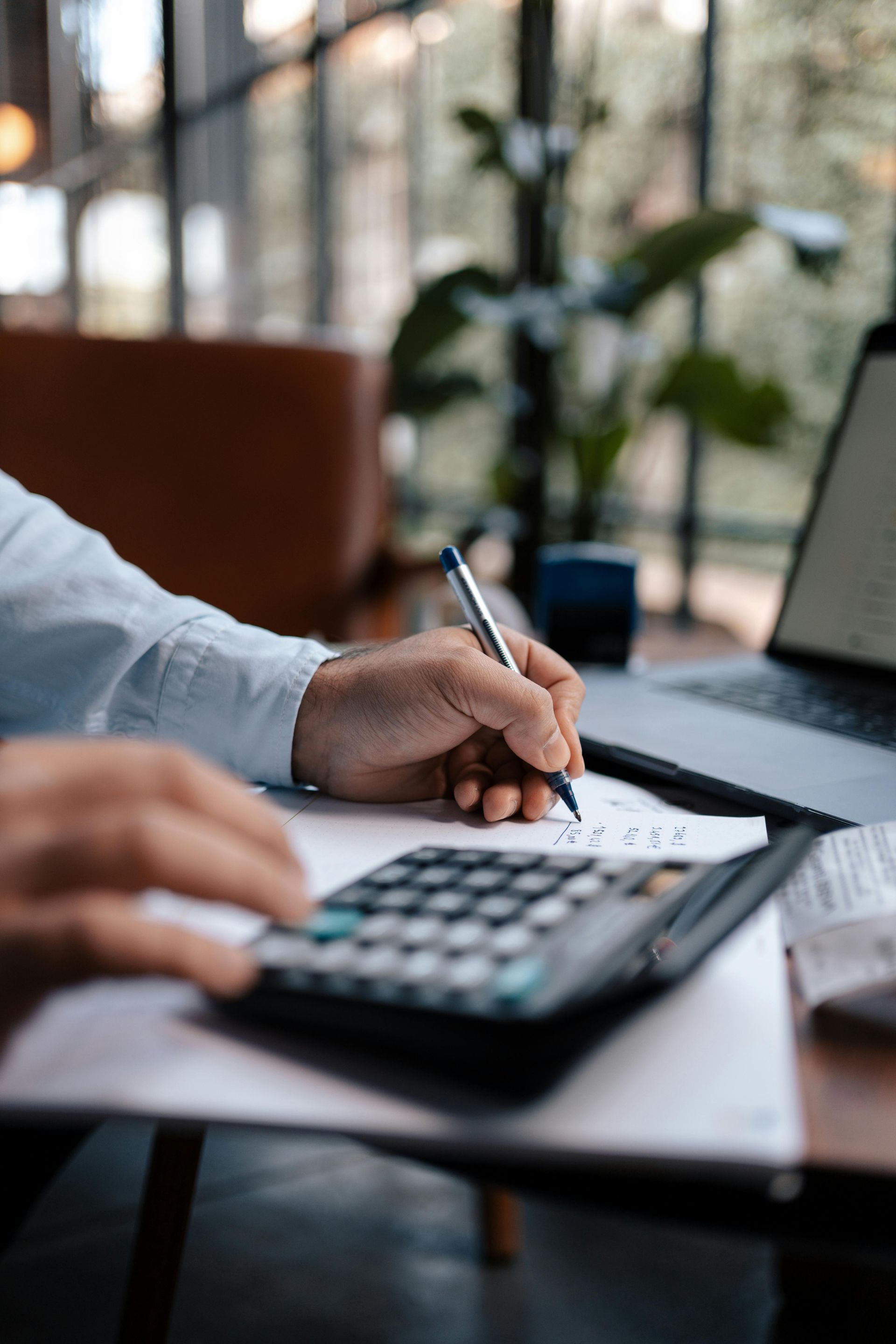 Person using a calculator and pen, working on paperwork next to a laptop by a window.