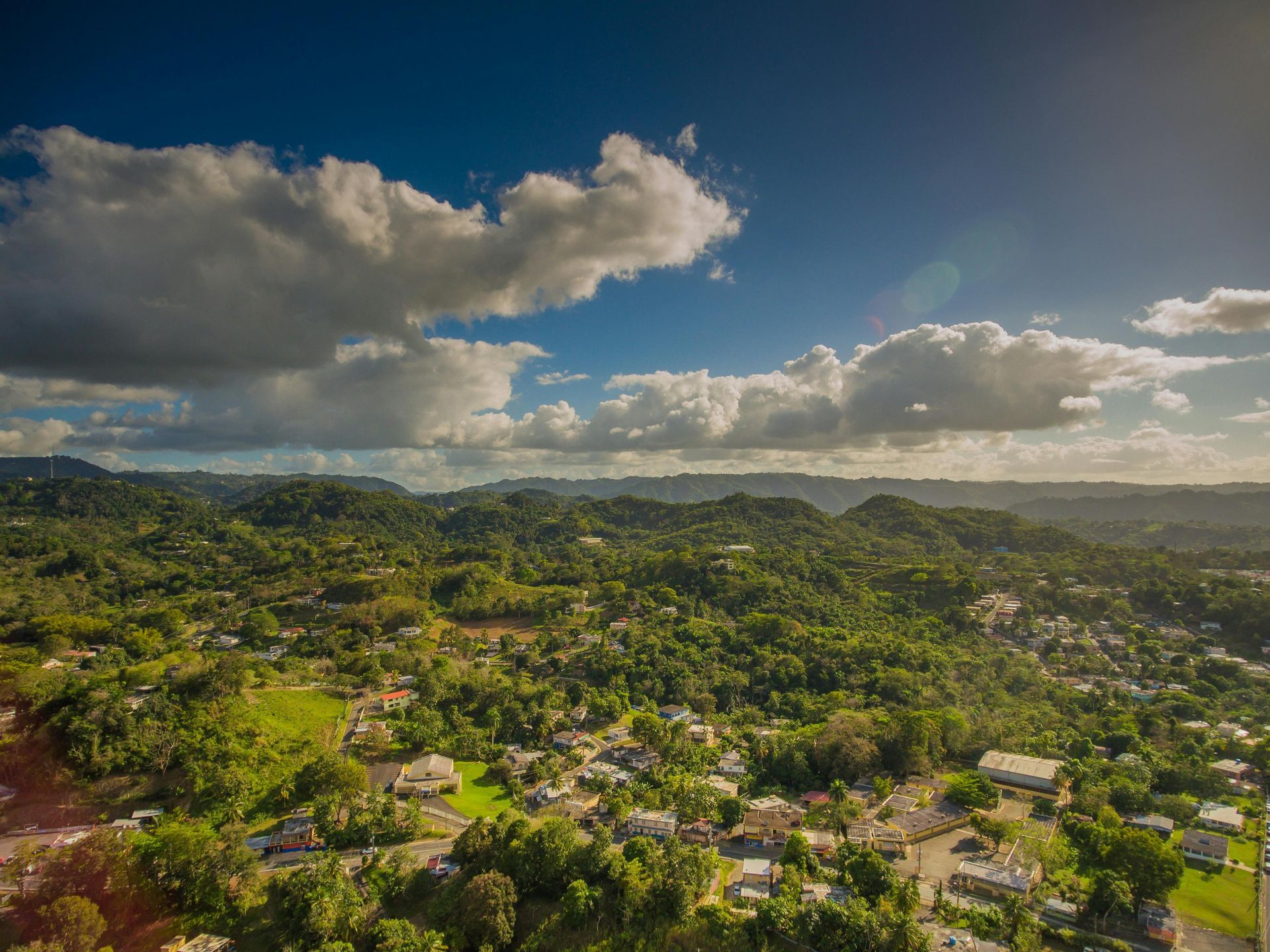 Green hills dotted with houses under a bright blue sky with fluffy white clouds.