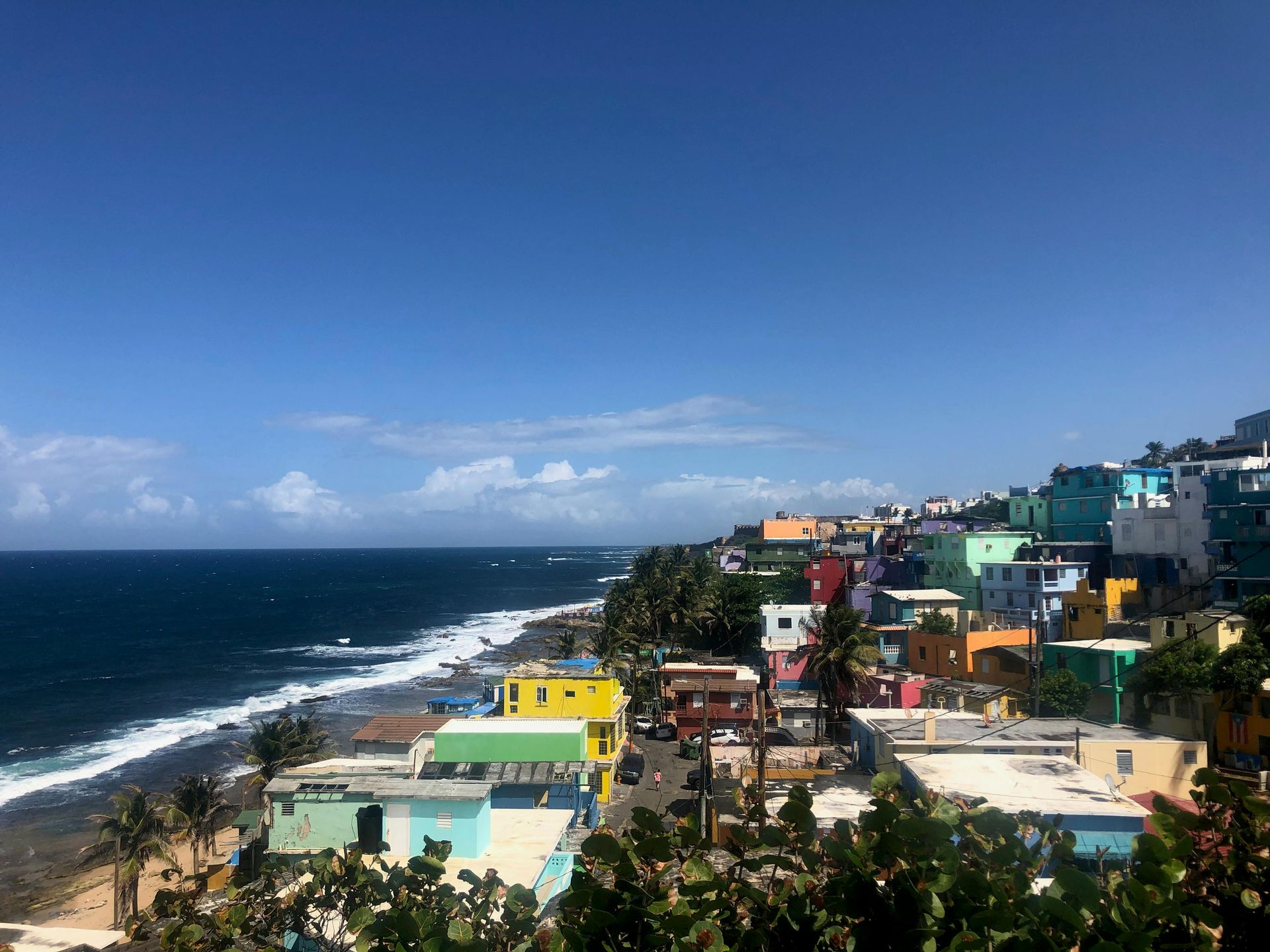 Colorful buildings line a coast under a bright blue sky, with ocean waves breaking in the foreground.
