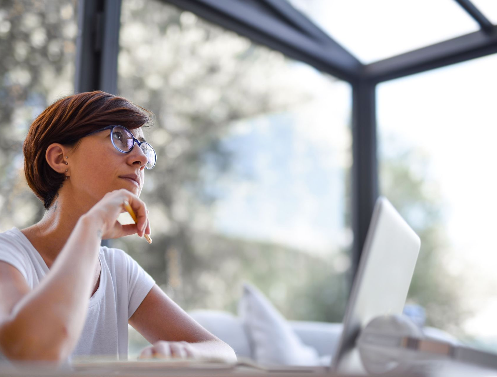 Person sitting at a table, thinking beside a laptop in a bright indoor workspace.