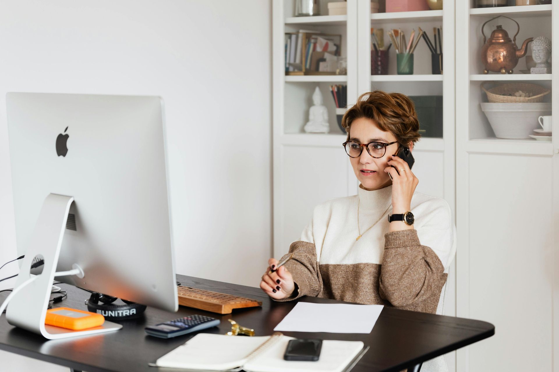 Woman on phone at a desk with computer, notepad, and calculator in a home office.