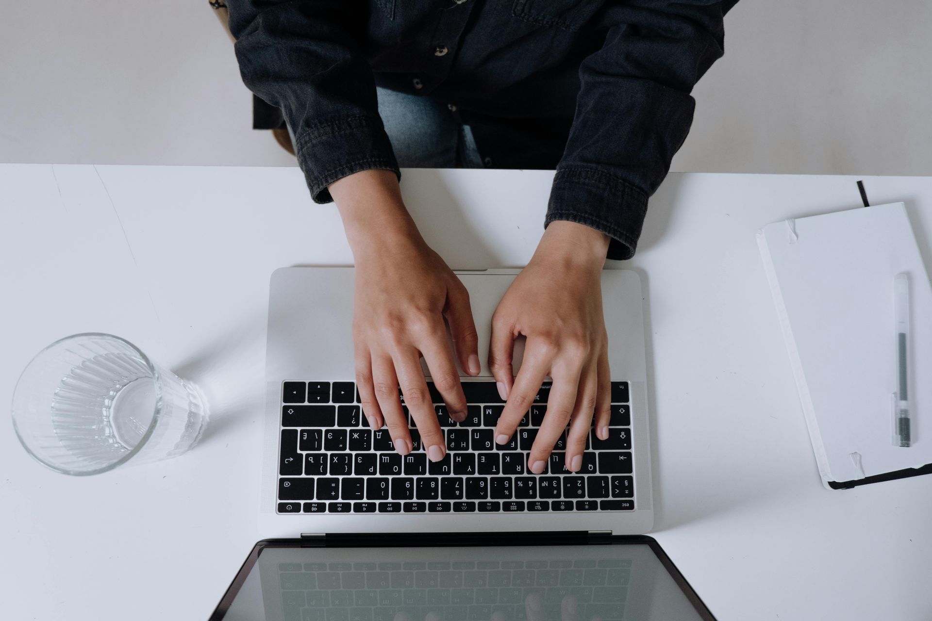Overhead view of hands typing on a laptop on a white desk with a glass of water and a notepad.