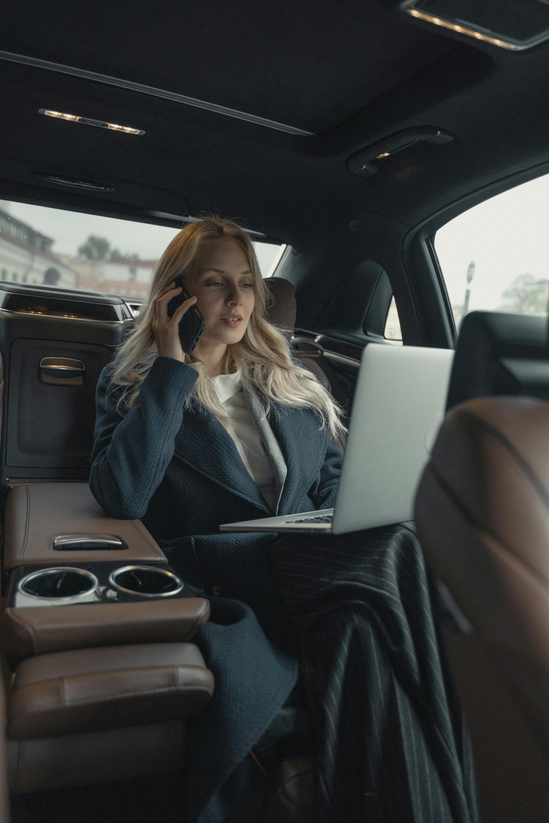 Woman in car on phone, using a laptop. Wearing a blue coat, sitting in a brown leather interior.