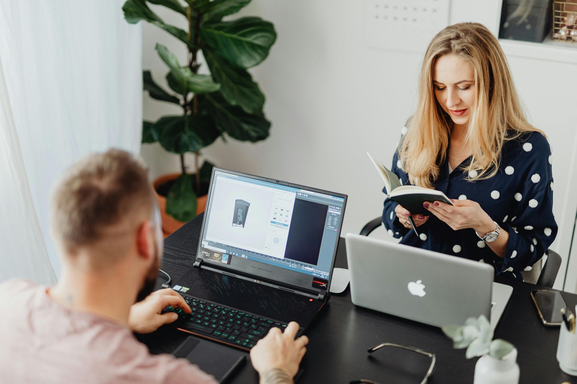 Two people working at a desk, one on a laptop with design software, the other looking at a notebook.