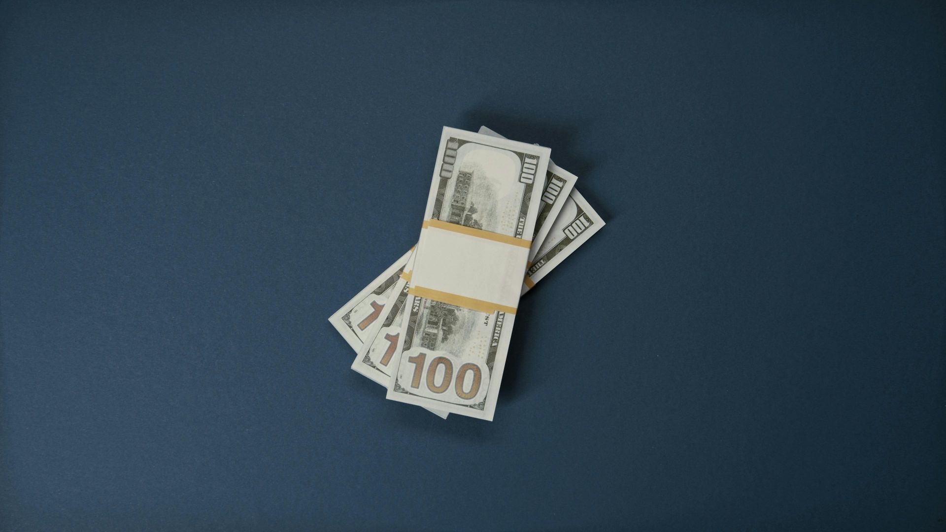 Stack of US $100 bills on a dark blue surface, bound with a tan paper band.