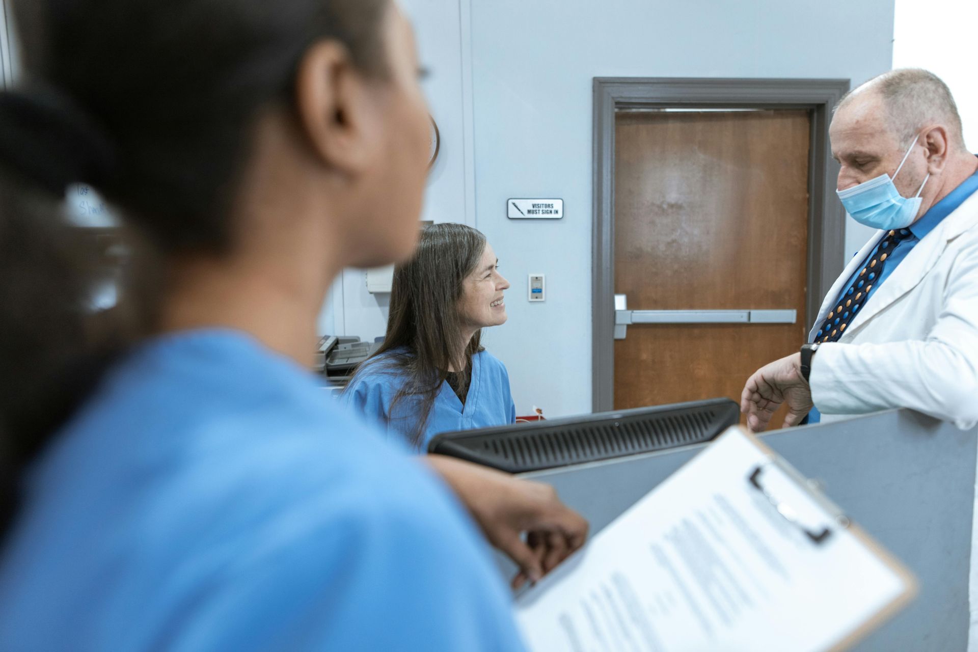Medical staff in scrubs and a lab coat converse at a reception desk, one wearing a face mask.