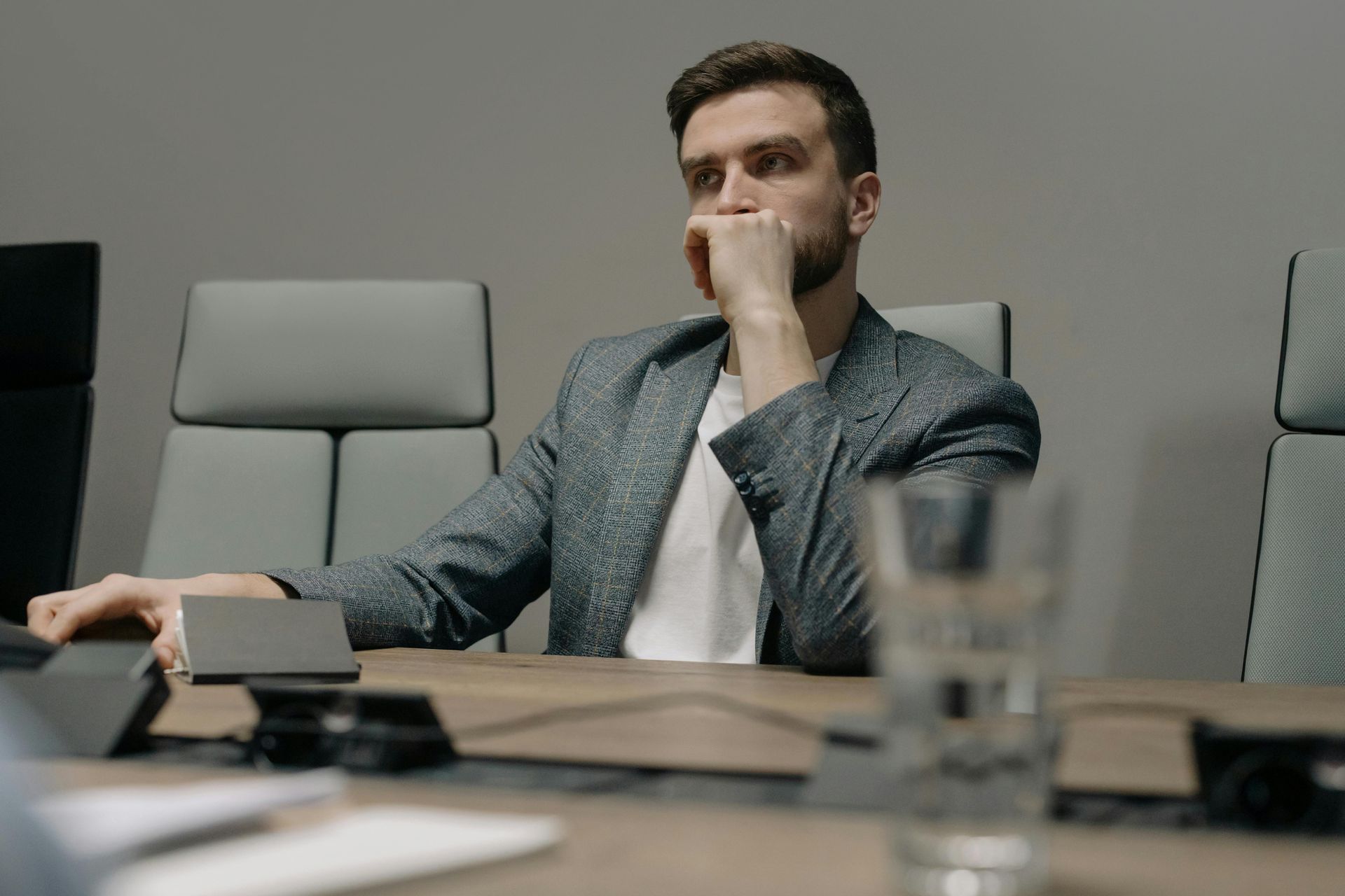 Man in a gray shirt sits at a conference table, resting his hand on his chin in a meeting room.
