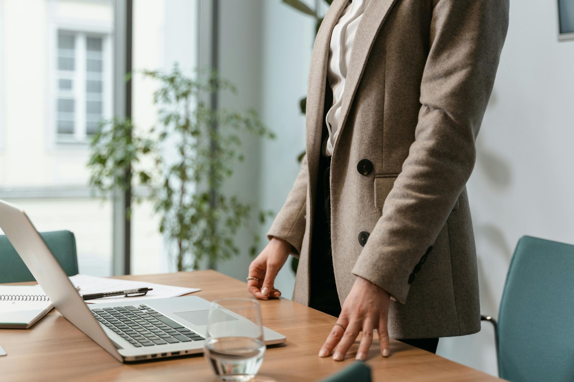 Person in blazer standing near a desk with laptop, glass of water, and chair in an office setting.