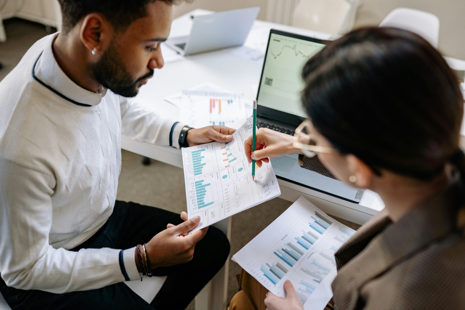 Two colleagues review printed charts at a desk with a laptop in a bright office.