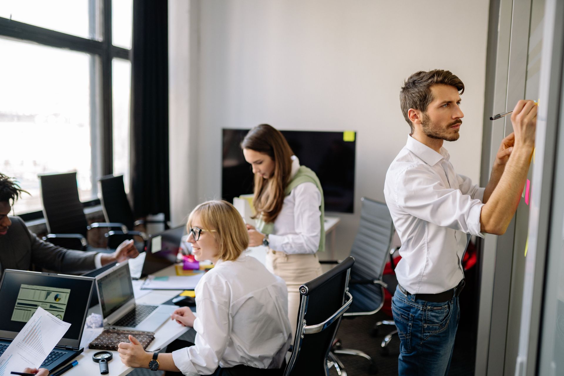 Team working in a modern office, one person writing on a glass board while others collaborate at desks.