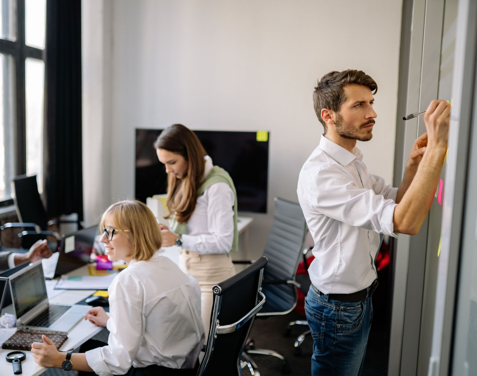 Three coworkers collaborating in a modern office, one writing on a glass wall, two working at desks