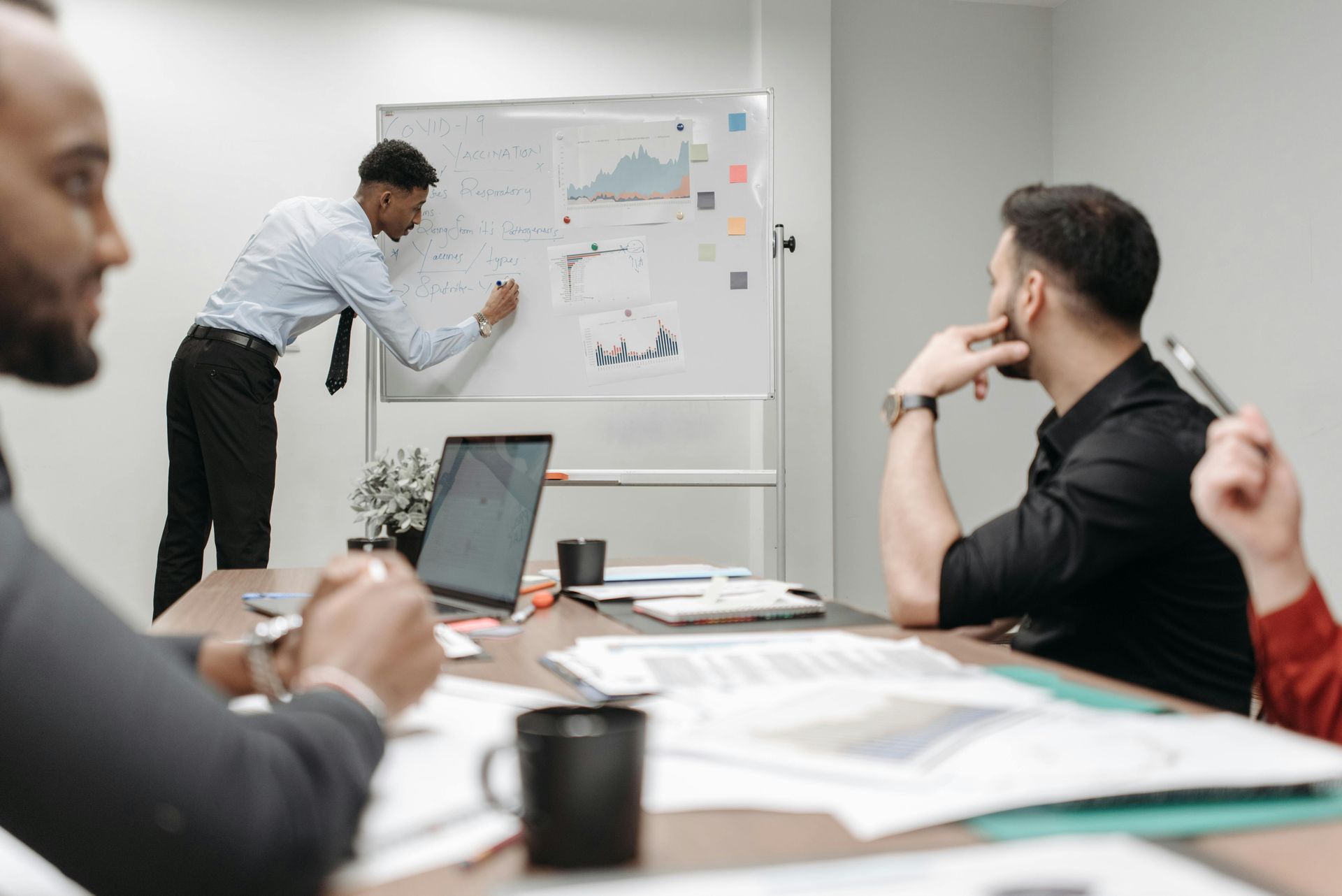A person stands at a whiteboard in an office setting, presenting data to colleagues seated at a table.