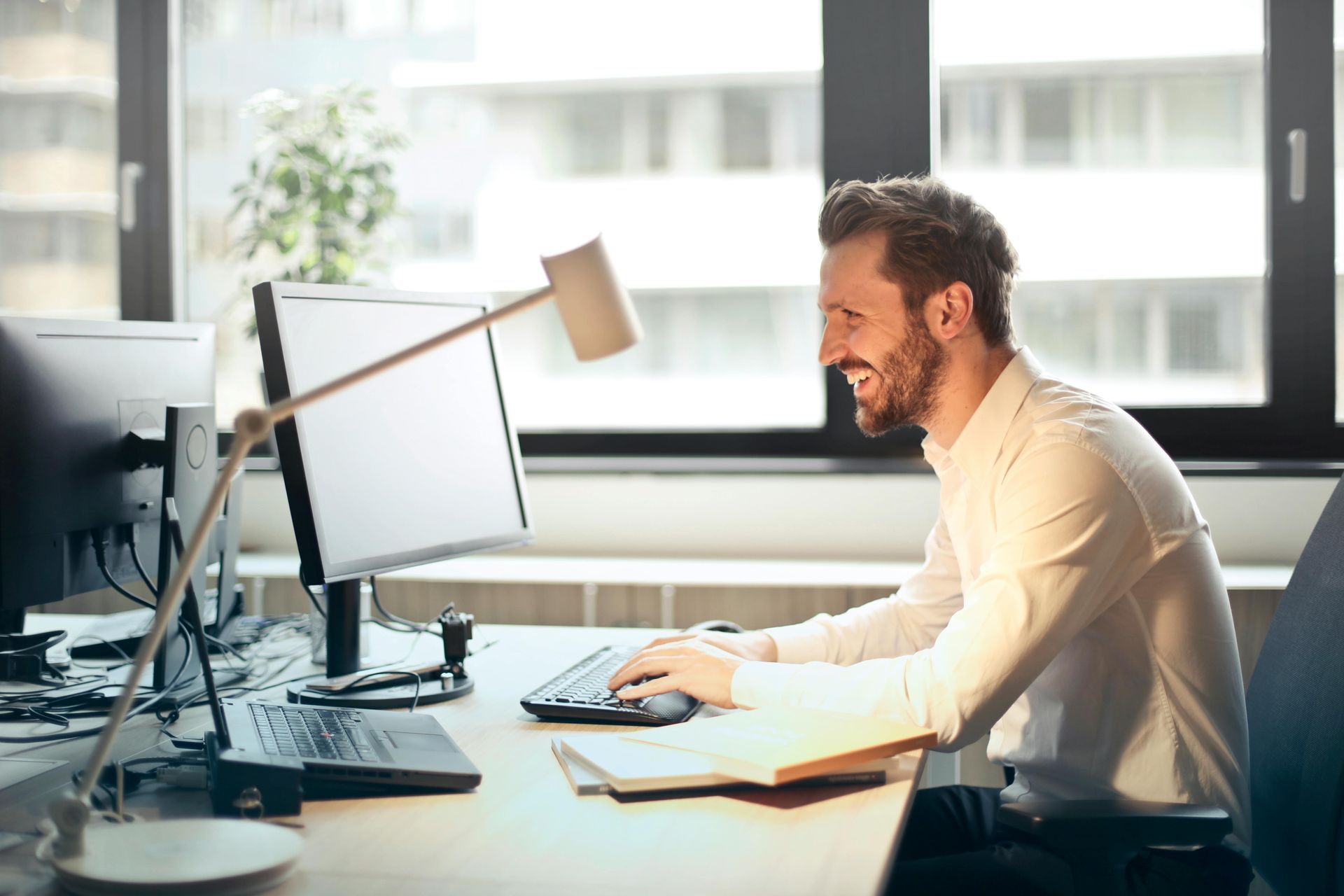 Man smiling while working at a computer in an office, desk with lamp and open book.