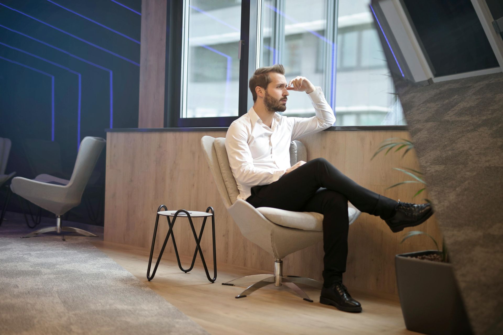 Man in white shirt and black pants, seated in a chair, looking out a window thoughtfully.