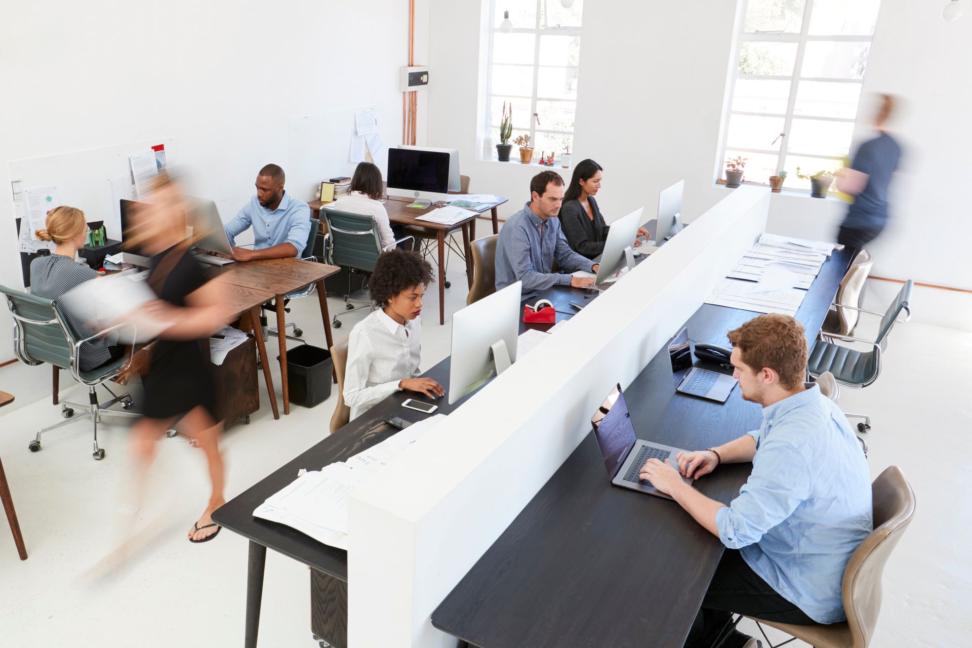 Open-plan office with people at desks, working on computers. Some are blurred as they move around.