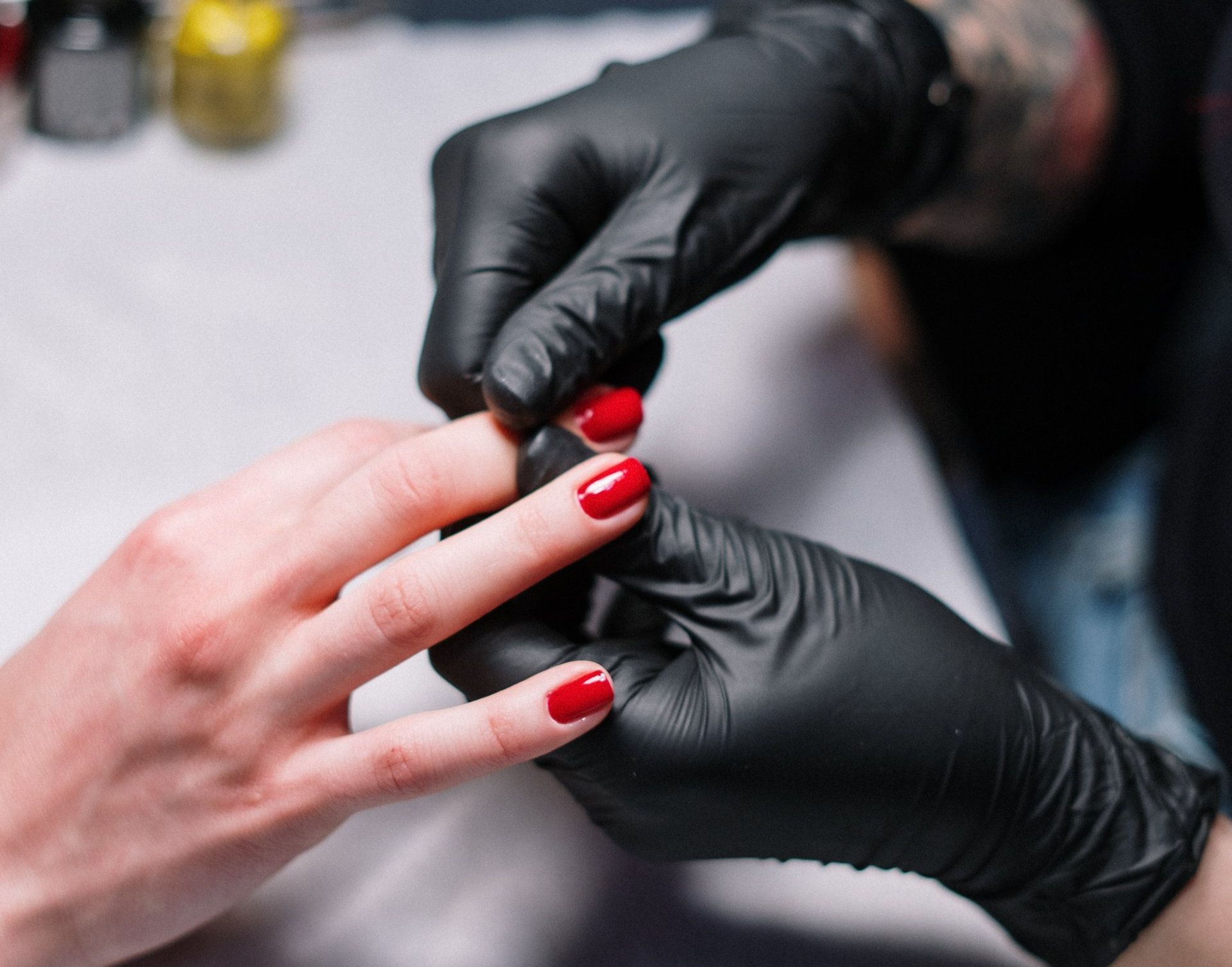 A gloved hand performs a manicure on a client's hand, which has vibrant red fingernails.
