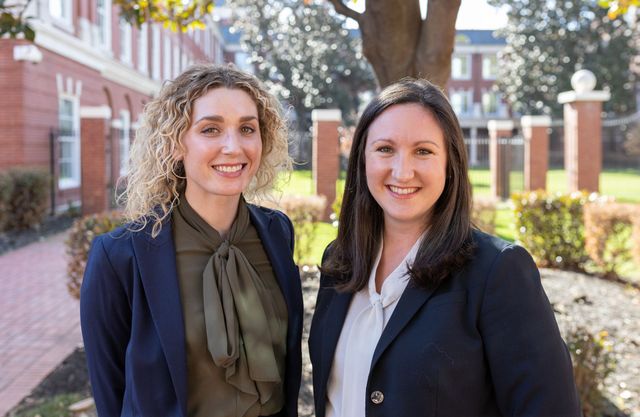 Two women smiling, posing outside a brick building, wearing business attire.