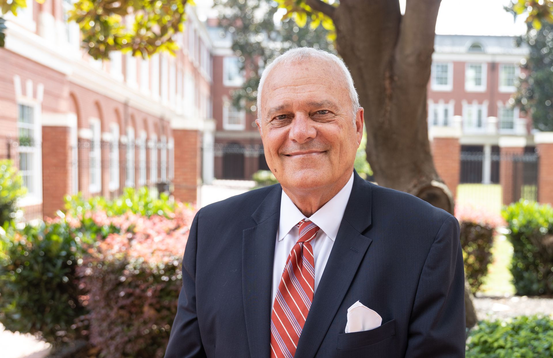 Man in a suit smiling, standing outdoors in front of a brick building.