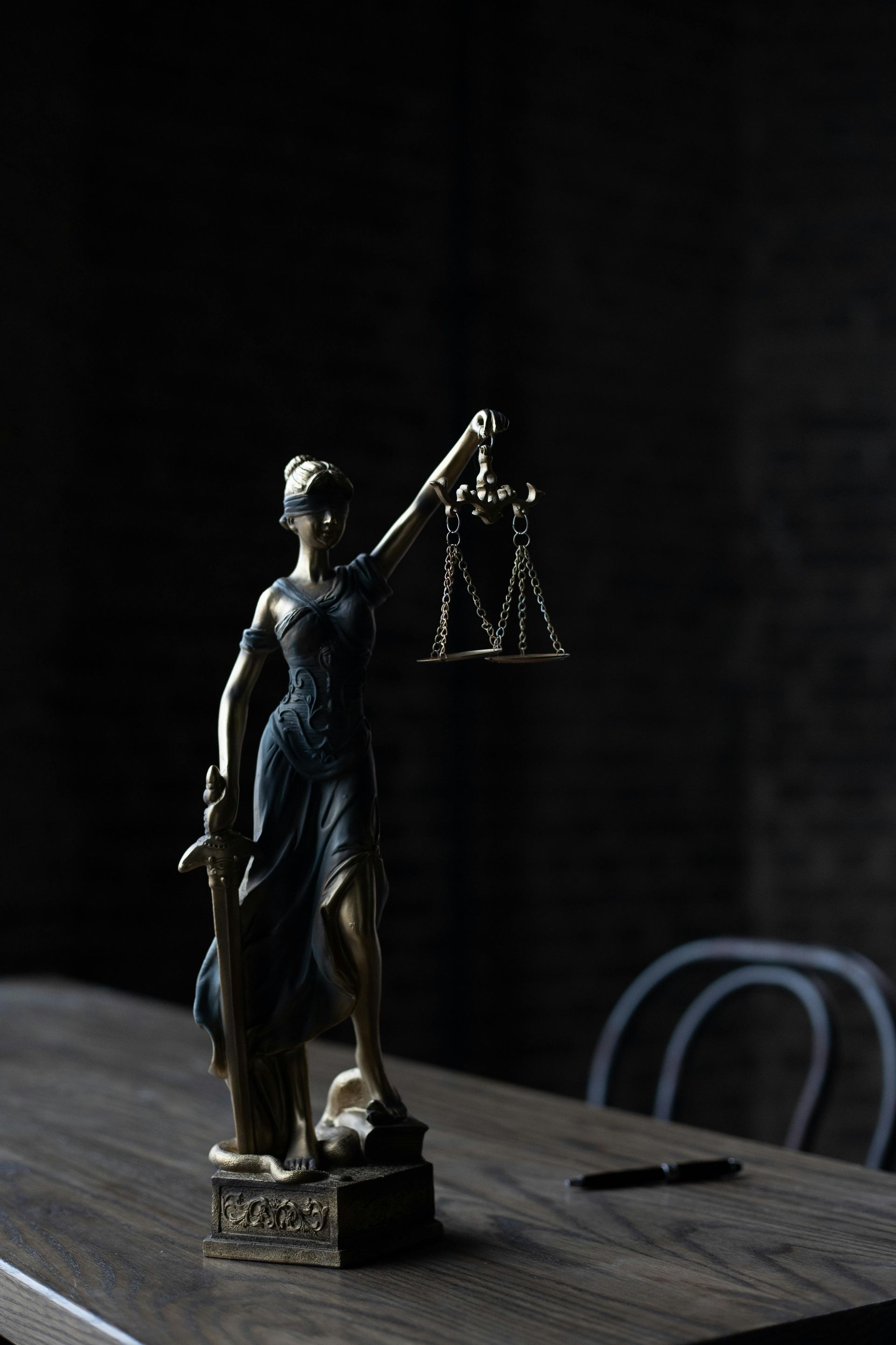 Statue of Lady Justice holding scales on a wooden table. Dark setting, with a pen and chair visible.