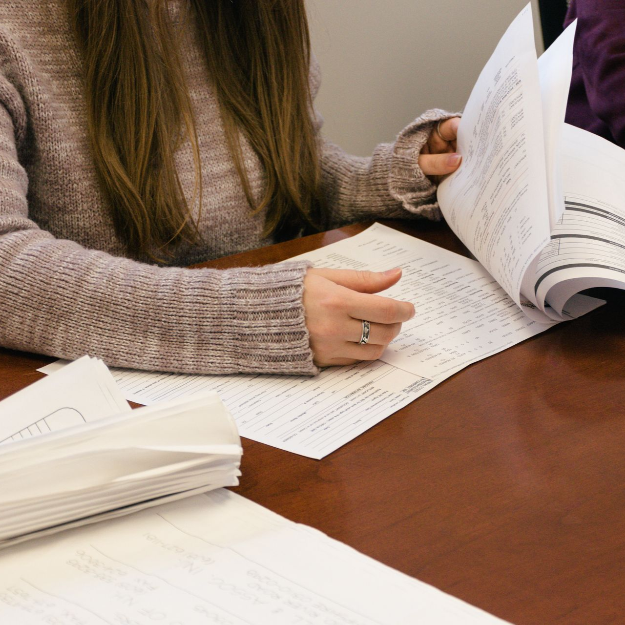 Two people at a table reviewing documents; one turns a page, papers spread around on a wooden surface.